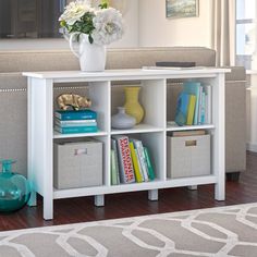 a white bookcase with baskets and flowers on it in front of a couch next to a window