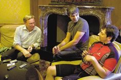 three men sitting on couches in front of a fire place holding remotes and looking at each other