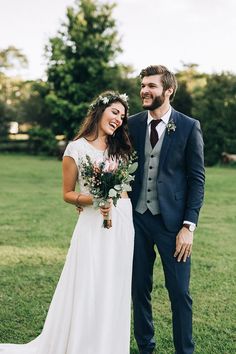 a bride and groom are standing in the grass with flowers on their head, smiling at each other