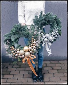 a woman holding a christmas wreath with ornaments on the front and sides, standing in front of a gray wall