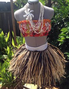 a dress made out of grass with beads and flowers on the top, sitting in front of some bushes