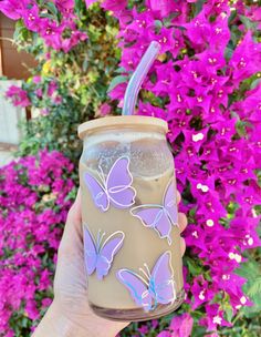 a hand holding a cup with a straw and butterfly decals on the side, in front of purple flowers