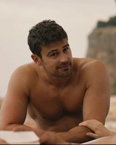 a shirtless man sitting on the beach looking at something in his hand while reading a book