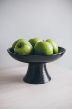 a black bowl filled with green apples sitting on top of a wooden table next to a white wall