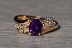 The Violet Town: Rich Amethyst and Diamond Ring in Yellow Gold. The ring centers on an oval brilliant cut natural rich purple amethyst. On either side of the center are two rows of round brilliant cut natural diamonds, one row of three, the other of two, that compliment the tapered bypass style shank. The ring is crafted in 14 karat yellow gold and is currently a finger size 5 yet can be adjusted to any finger size for an additional charge on request. The ring is comfortable to wear and features Oval Purple Diamond Ring With Gemstone Accents, Purple Amethyst Oval Diamond Ring, Oval Amethyst Cubic Zirconia Ring, Oval Amethyst Ring With Cubic Zirconia, Oval Purple Amethyst Ring With Cubic Zirconia, Purple Oval Brilliant Cut Ring, Oval Purple Diamond Ring With Center Stone, Dazzling Purple Oval Rings, Formal Oval Amethyst Ring With Cubic Zirconia