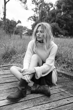 a woman sitting on top of a wooden floor next to tall grass and trees in the background