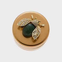 a gold and green bee brooch with diamonds on it's back, sitting on top of a round box