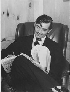 a man in a suit and tie sitting on a chair reading a paper while holding a pen