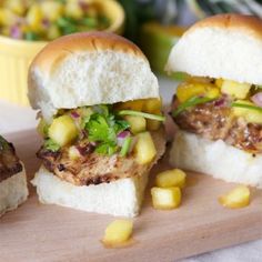 three sandwiches with meat, pineapple salsa and onions on a cutting board next to a bowl of corn