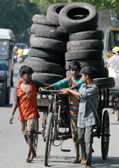 two boys pushing a cart with tires on it down the street while another boy pushes a bicycle behind them