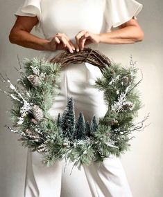 a woman is holding a wreath with pine cones and evergreens on it, while standing in front of a white background