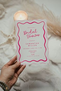 a person holding up a pink and white bridal shower card next to a lit candle