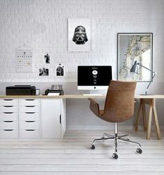 a desk with two computers on it in front of a white brick wall and wooden chairs