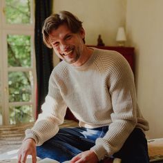 a smiling man sitting on top of a bed next to a window and holding a book