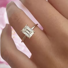 a woman's hand with a diamond ring on top of her finger and pink flowers in the background