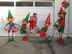 three garden gnomes are holding gifts in front of a garage door and one is carrying a present