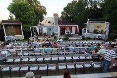 an outdoor stage with people sitting on chairs and facing the stage, in front of several houses