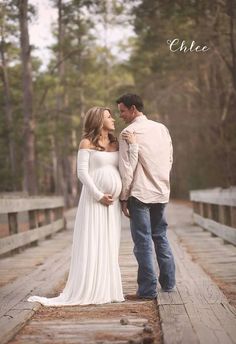 a pregnant couple standing on a bridge in the woods with their arms around each other