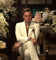 a man in a white suit and tie sitting next to a table with cakes on it