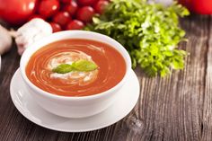 a bowl of tomato soup on a wooden table next to fresh tomatoes and parsley