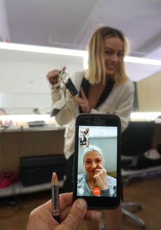 a woman is taking a selfie with her cell phone in an office setting, while another woman stands behind her