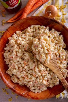 a wooden bowl filled with macaroni salad next to carrots and an onion