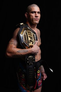 a man holding a wrestling belt in his right hand and looking at the camera while standing against a black background