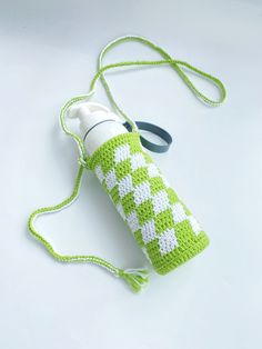 a green and white crocheted water bottle cover with a black handle on a white surface