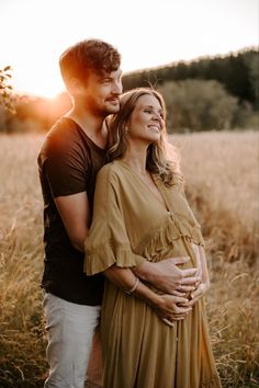 a pregnant couple standing in the middle of a field at sunset with their arms around each other