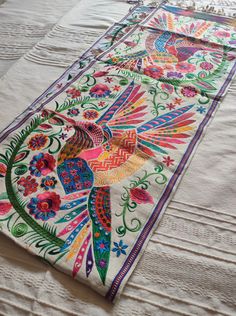 an embroidered table runner with colorful birds and flowers on white fabric, sitting on top of a bed