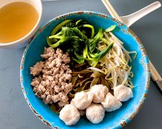 a blue bowl filled with meat and veggies next to a cup of tea