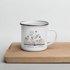 a white enamel mug with an open book and flowers on it sitting on a wooden tray