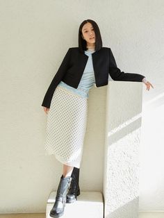 a woman leaning against a wall with her hands on her hips, wearing a black jacket and white polka dot skirt