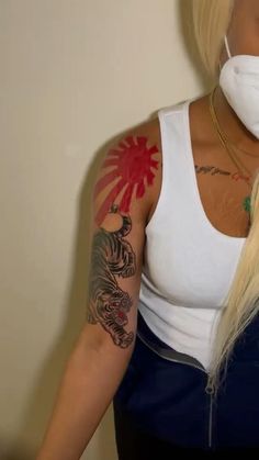 a woman wearing a face mask with tattoos on her arms and shoulder, standing in front of a wall