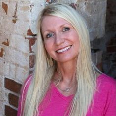 a woman with long blonde hair standing in front of a brick wall smiling at the camera