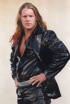 a man with long hair wearing a black leather jacket and silver bracelets standing in front of a white wall