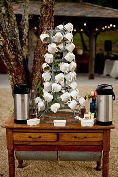 a wooden table topped with lots of white cups and saucers next to a tree