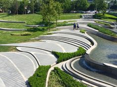 two people are walking on the path next to an artificial water feature in a park