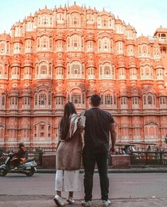 a man and woman standing in front of an ornate building with motorcycles passing by on the street