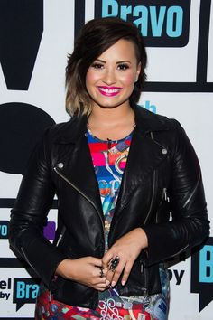 a woman wearing a black leather jacket and colorful shirt smiles at the camera with her hands in her pockets