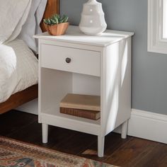 a white nightstand with a book on it next to a bed and a plant in a vase