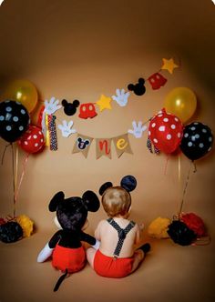 a baby sitting on the floor with mickey and minnie mouse balloons in front of it