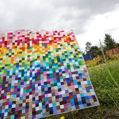 a multicolored quilt sitting on top of a lush green field next to tall yellow flowers