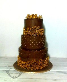 a three tiered chocolate cake with gold decorations on the top and bottom, sitting on a wooden table