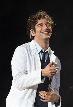 a man with curly hair wearing a white jacket and tie laughing while holding a microphone