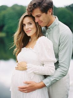 a pregnant couple standing next to each other in front of a body of water and trees