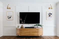 Flat screen TV hung above curved walnut cabinet symmetrically framed by botanical prints and gold arm sconces create the focal point for this room. White walls with chair rail and architectural trim contrast with hardwood flooring for a clean and beautiful traditional look. Art On Sides Of Tv, Moulding On Tv Wall, Molding Behind Tv, Big Wall With Tv Decor Living Room, Blank Wall Tv Ideas, Tv Wall With Molding, Living Room Mounted Tv Ideas, Box Trim Tv Wall, Photos Around Tv On Wall