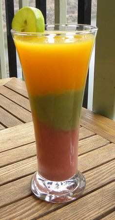 an orange, green and red drink sitting on top of a wooden table next to a slice of apple