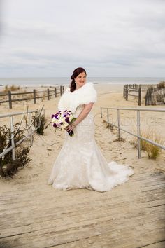 a woman in a wedding dress standing on a boardwalk near the beach with her bouquet