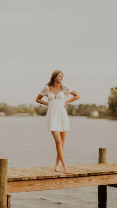 a woman in a white dress standing on a dock next to the water with her hands on her hips